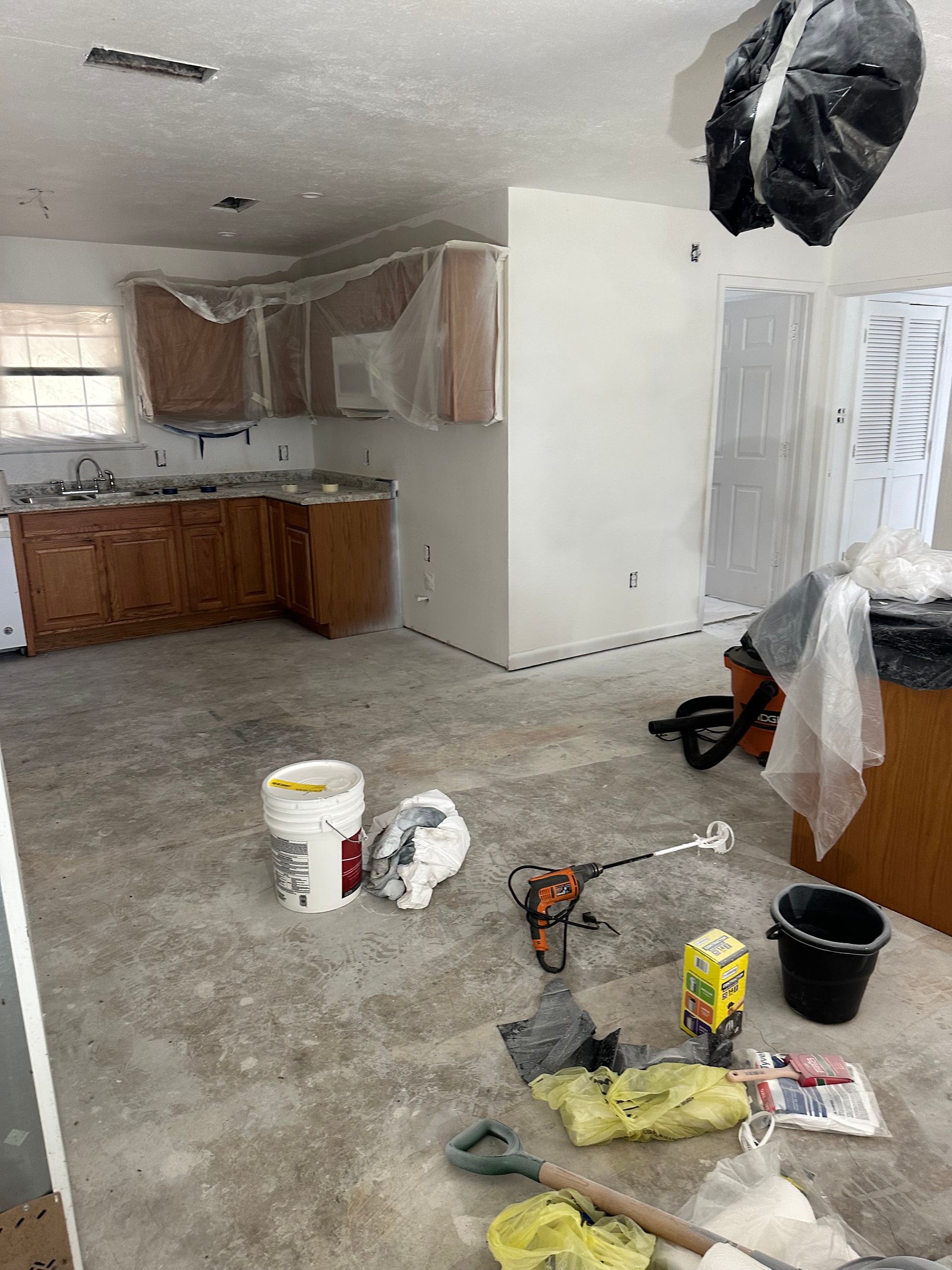 Partially renovated kitchen with bare floors, covered cabinets, paint buckets, tools, and construction debris