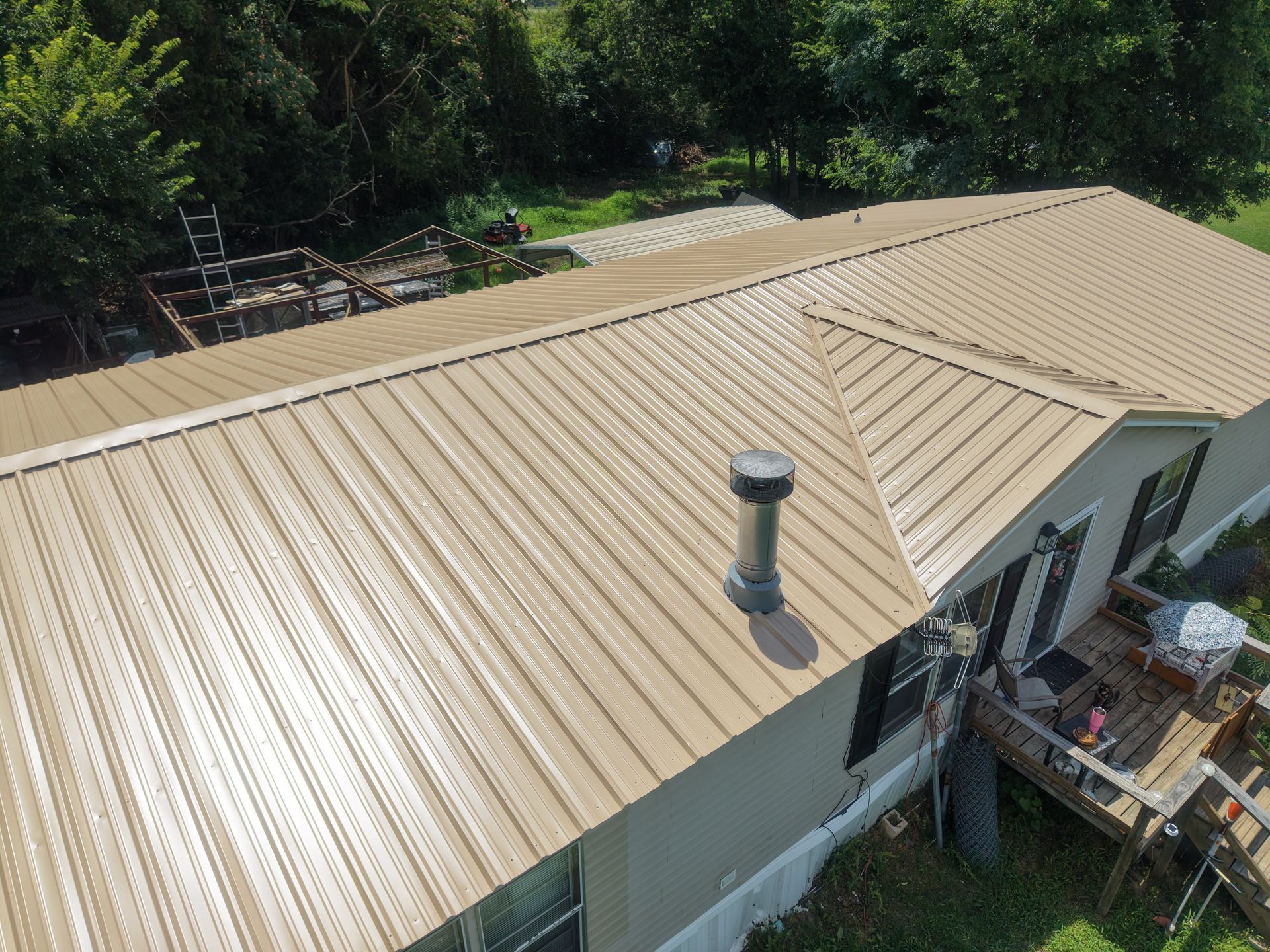 Tan metal roof with a vent pipe on a house, viewed from above, with trees in the background