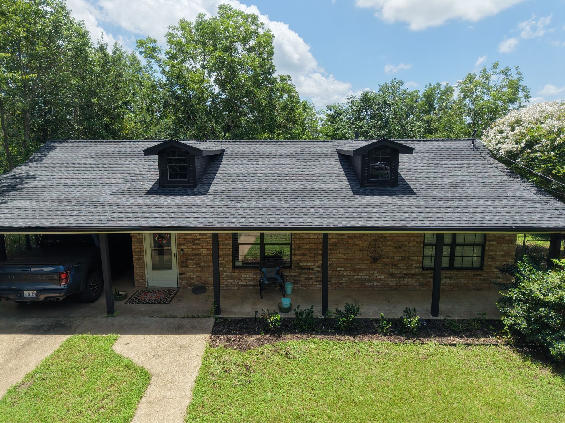 Brick house with gray roof, two dormers, front porch, and carport, surrounded by trees and lawn