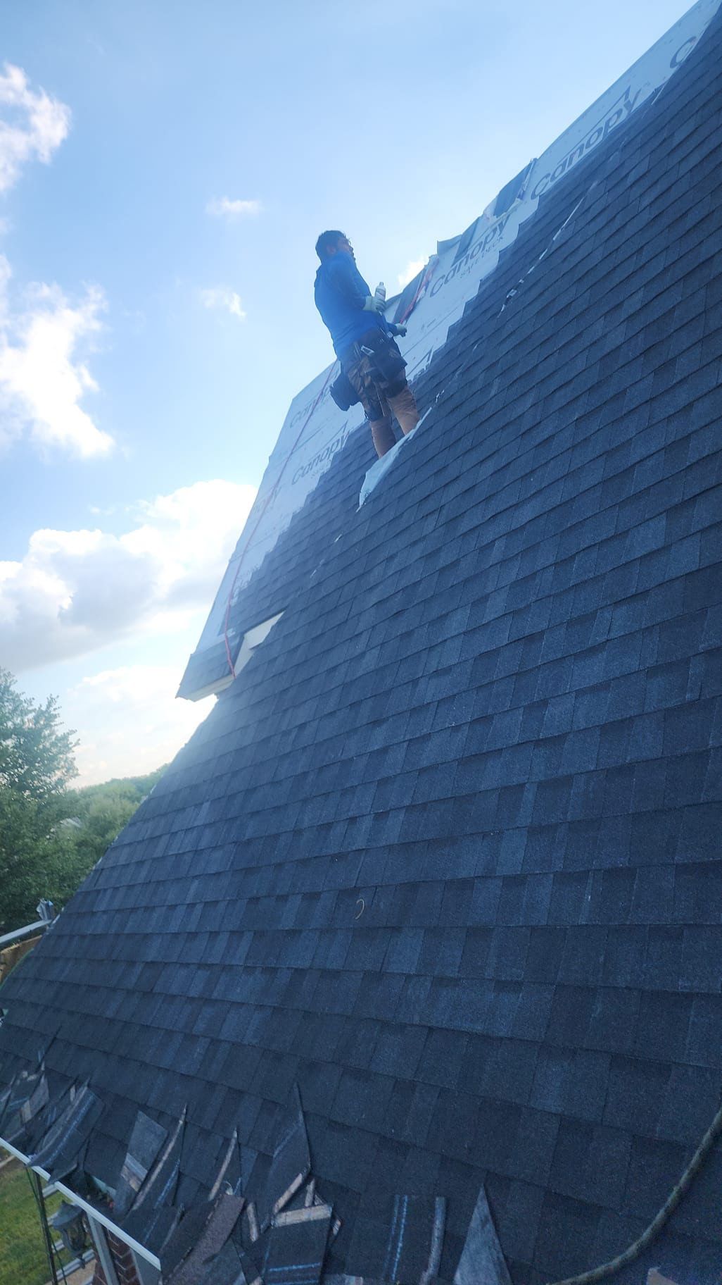 Person climbing a steep dark shingled roof under a blue sky.