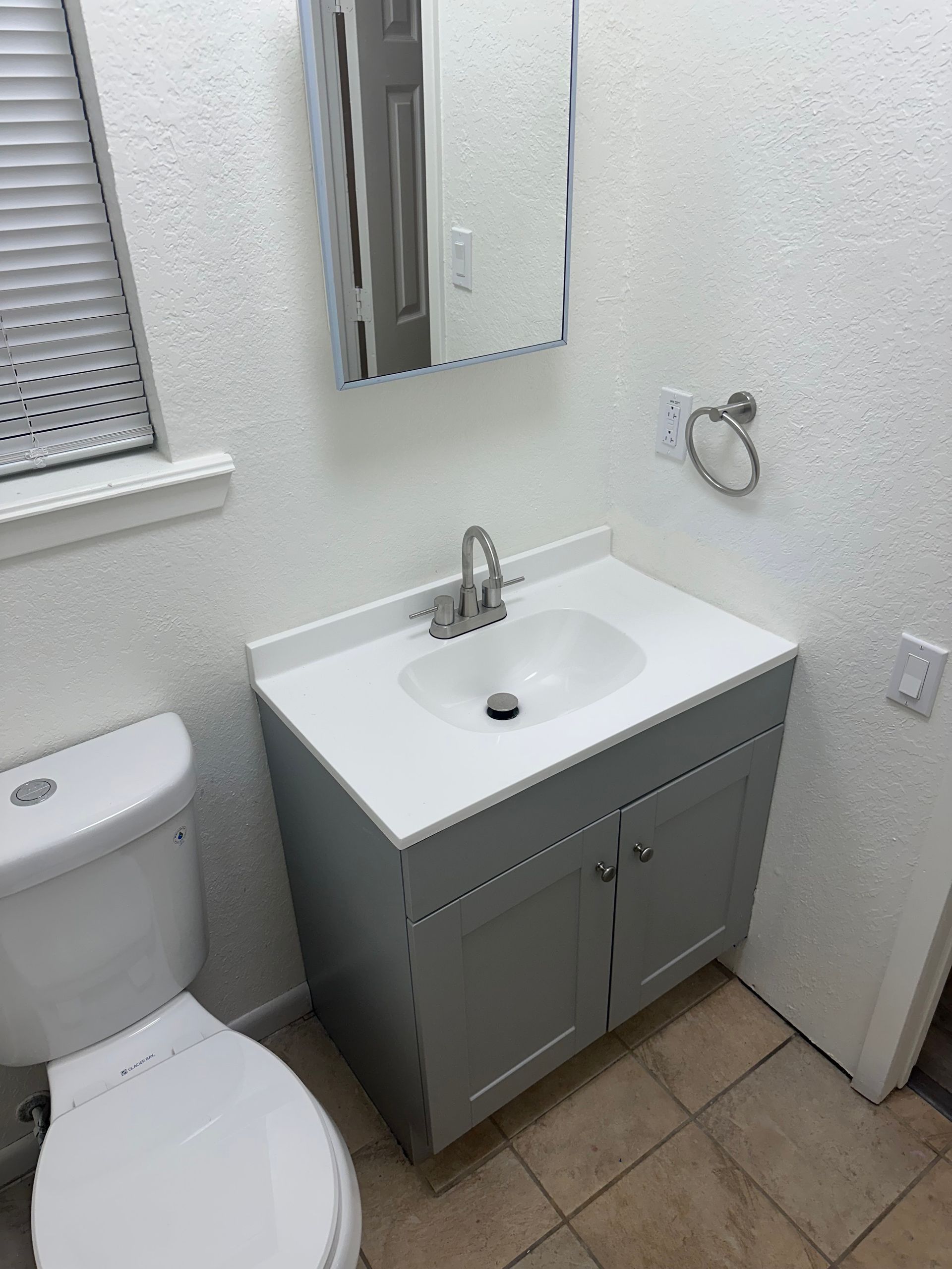 Small bathroom with white sink, gray vanity, toilet, mirror, and beige tile floor.