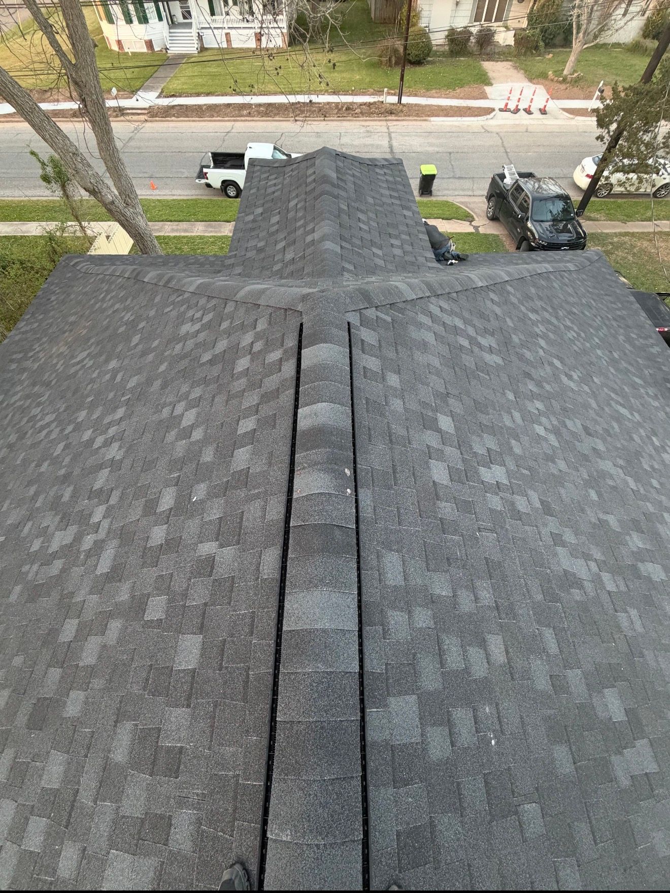 View from a roof over asphalt shingles to a street with parked trucks and leafless trees