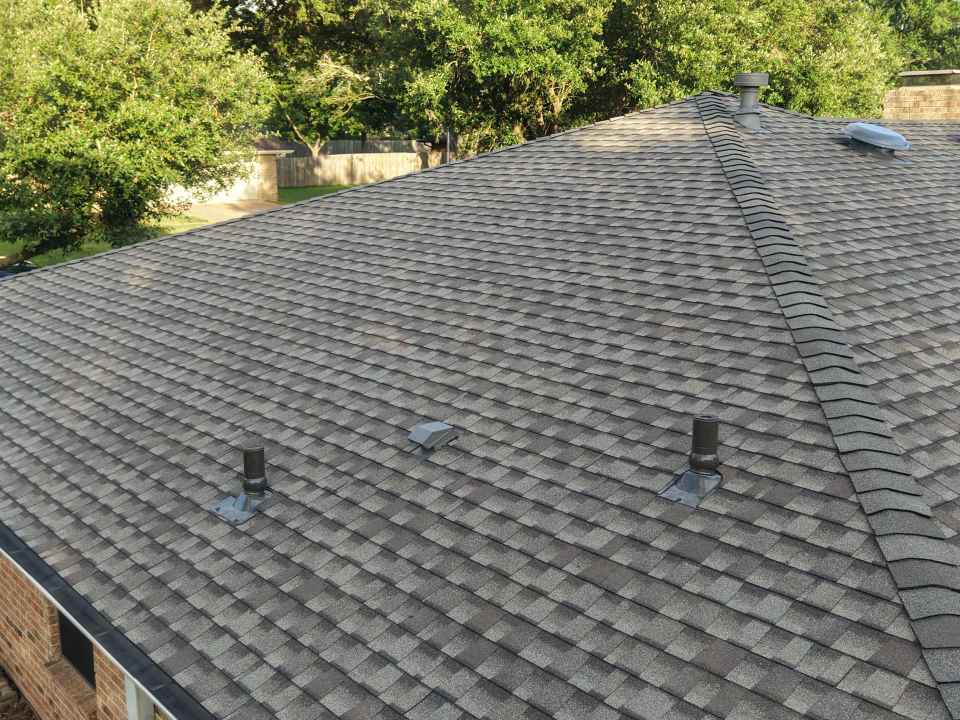 Gray shingled roof with several vents, surrounded by green trees and viewed from above