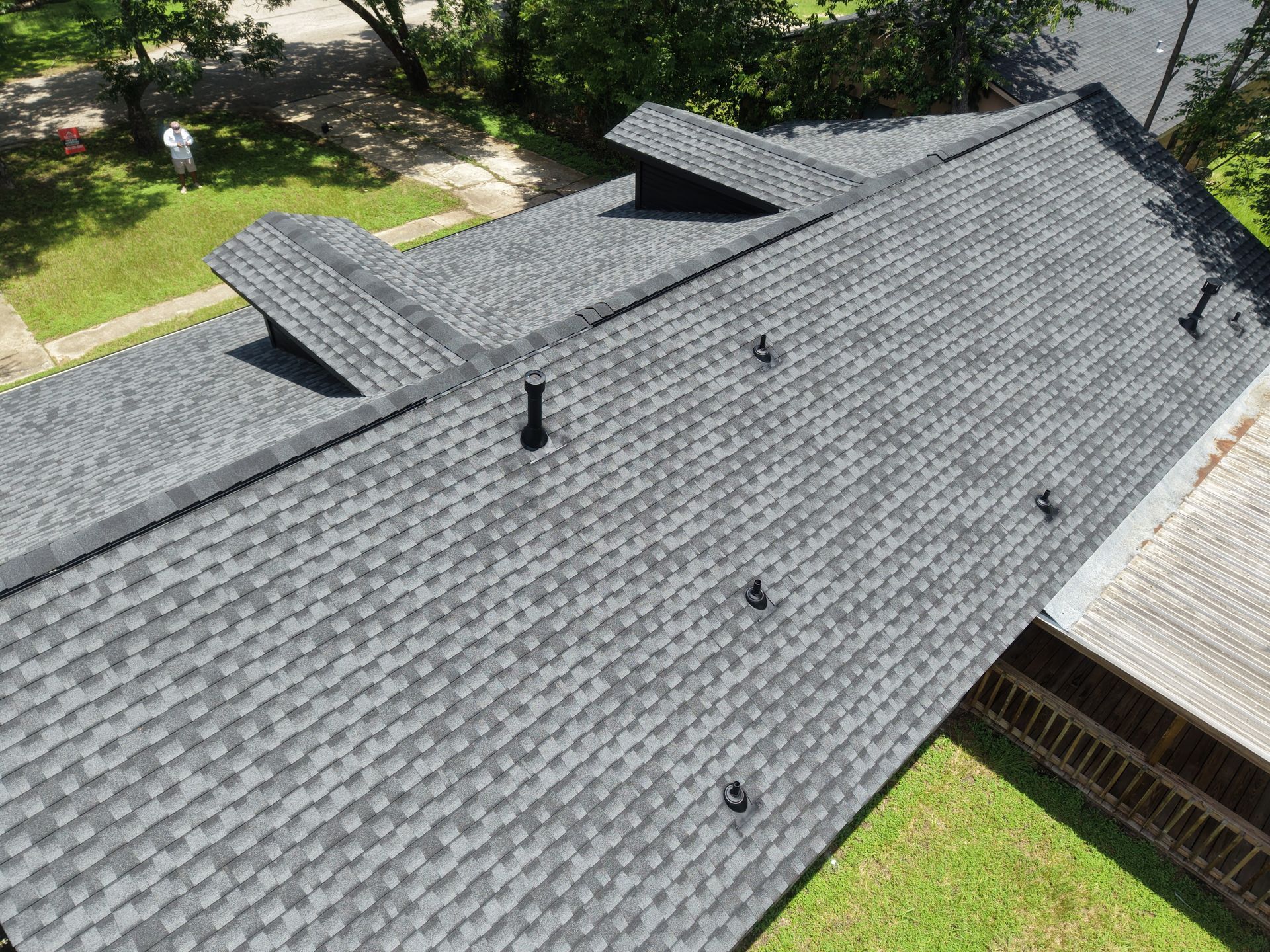 Aerial view of a gray shingled roof with multiple dormers and a chimney beside a green yard