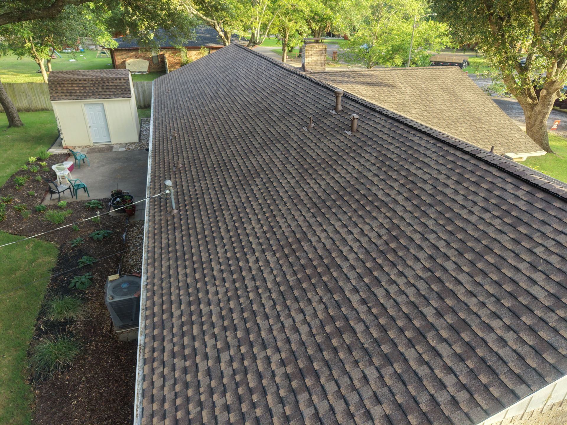 Aerial view of a house roof with brown shingles, trees, and yard below.