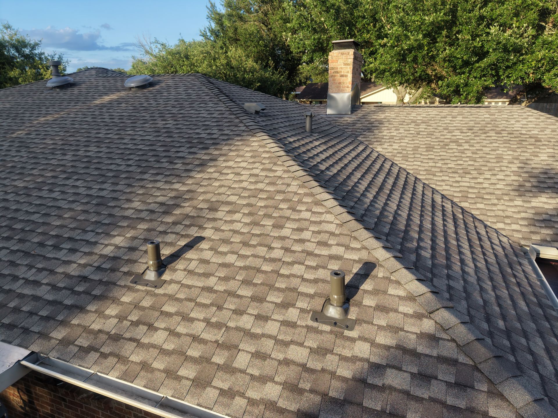 Rooftop with gray shingles, chimney, and two small roof vents under trees