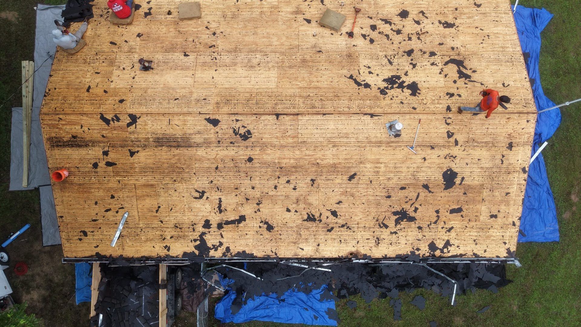 Top view of a weathered plywood roof with scattered debris and blue tarp edges around it