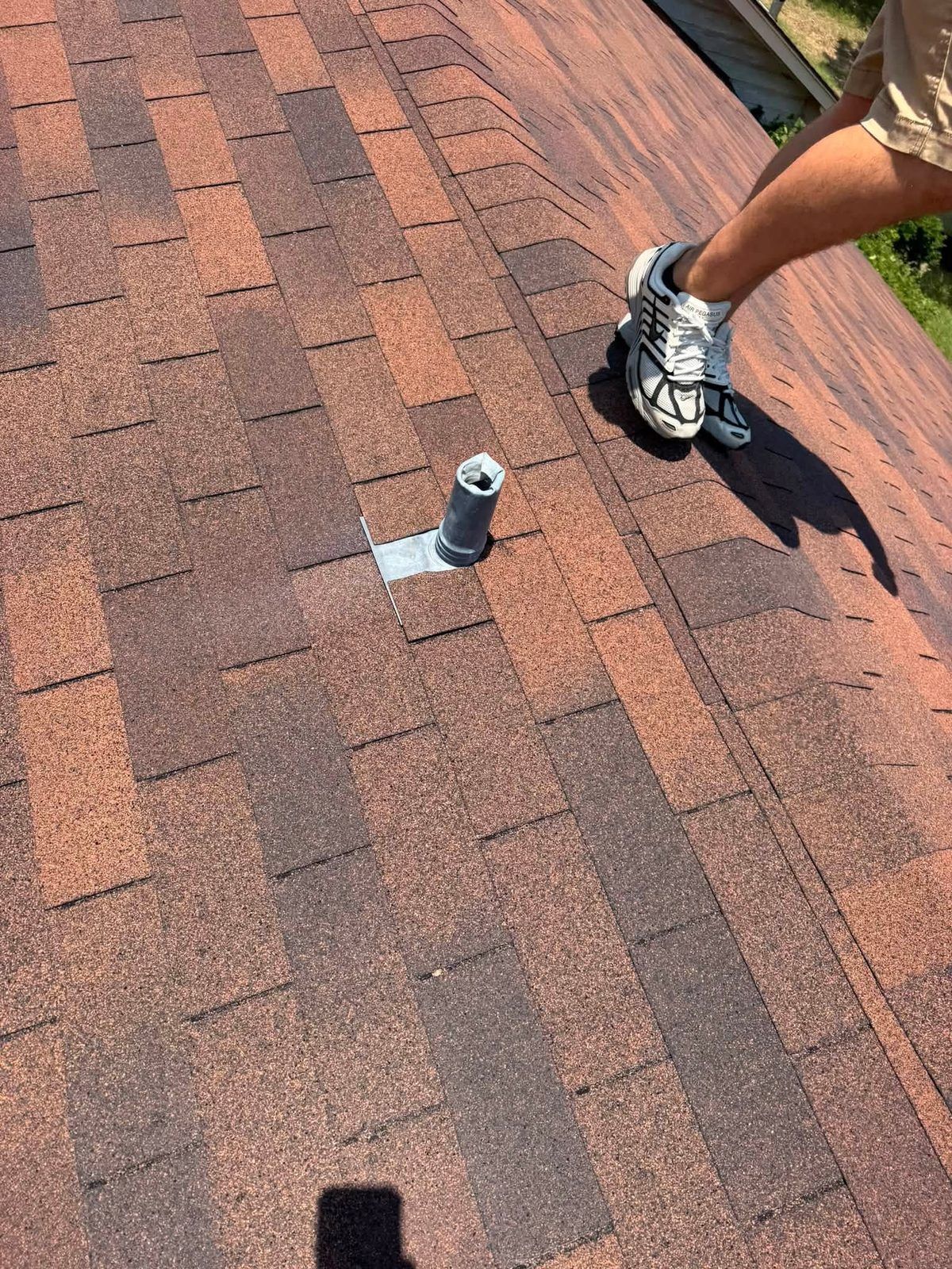 Roof shingles with a vent pipe opening and a worker’s boot on the right edge.