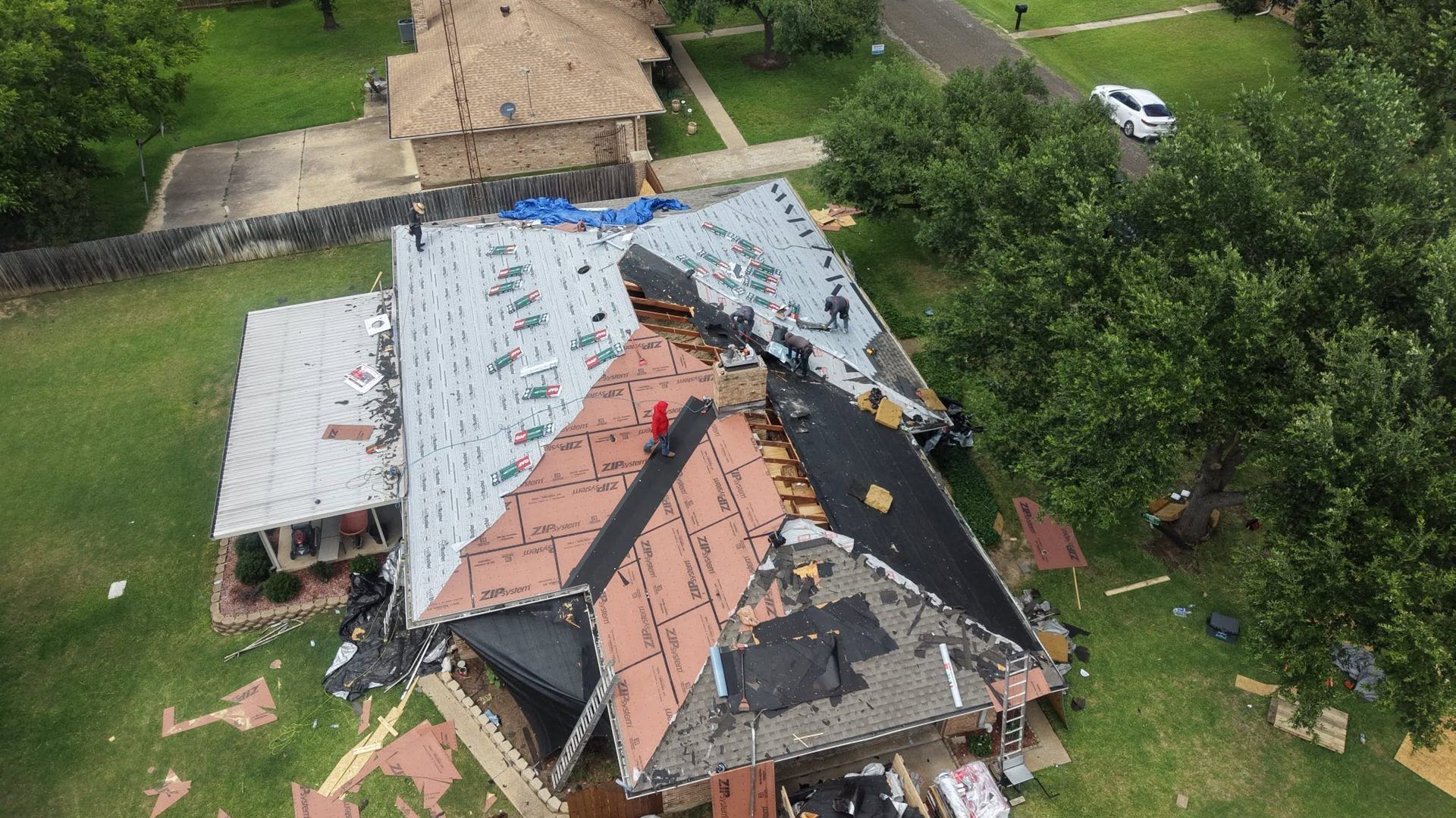 Aerial view of a house with a damaged roof and missing shingles, surrounded by trees and lawn.