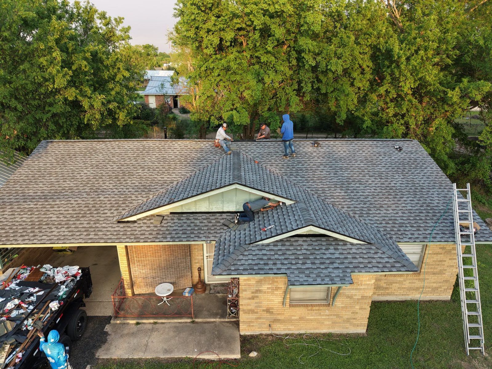 Aerial view of a house roof with two workers and a ladder near trees and parked cars