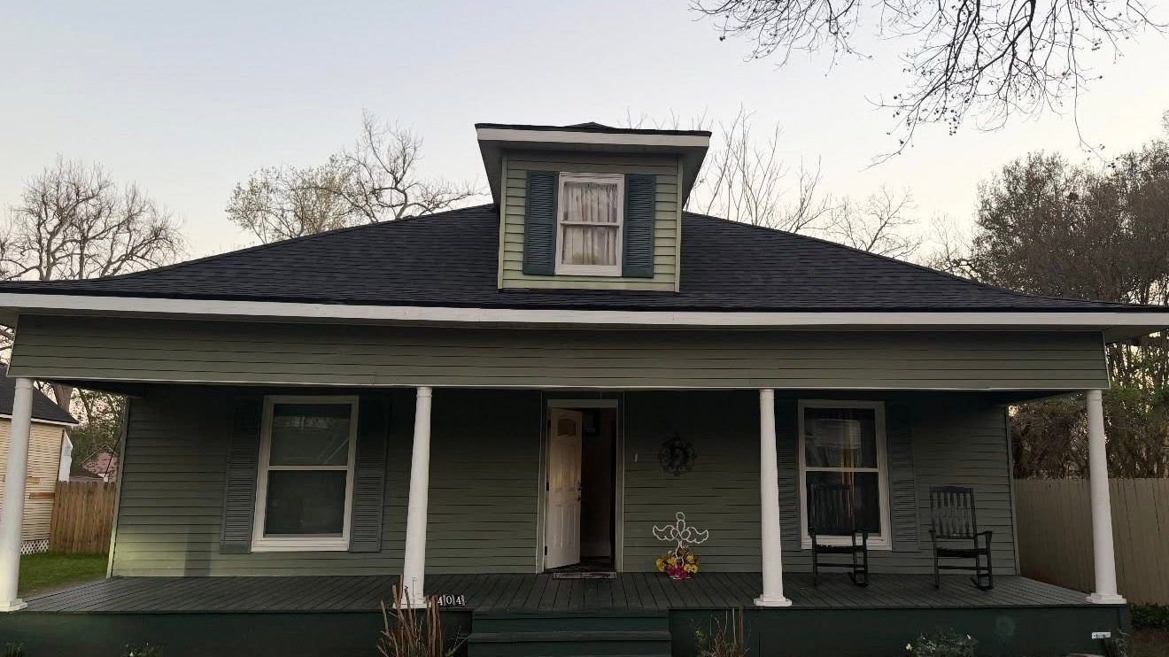 Small green house with a covered porch and central dormer, viewed from the front.