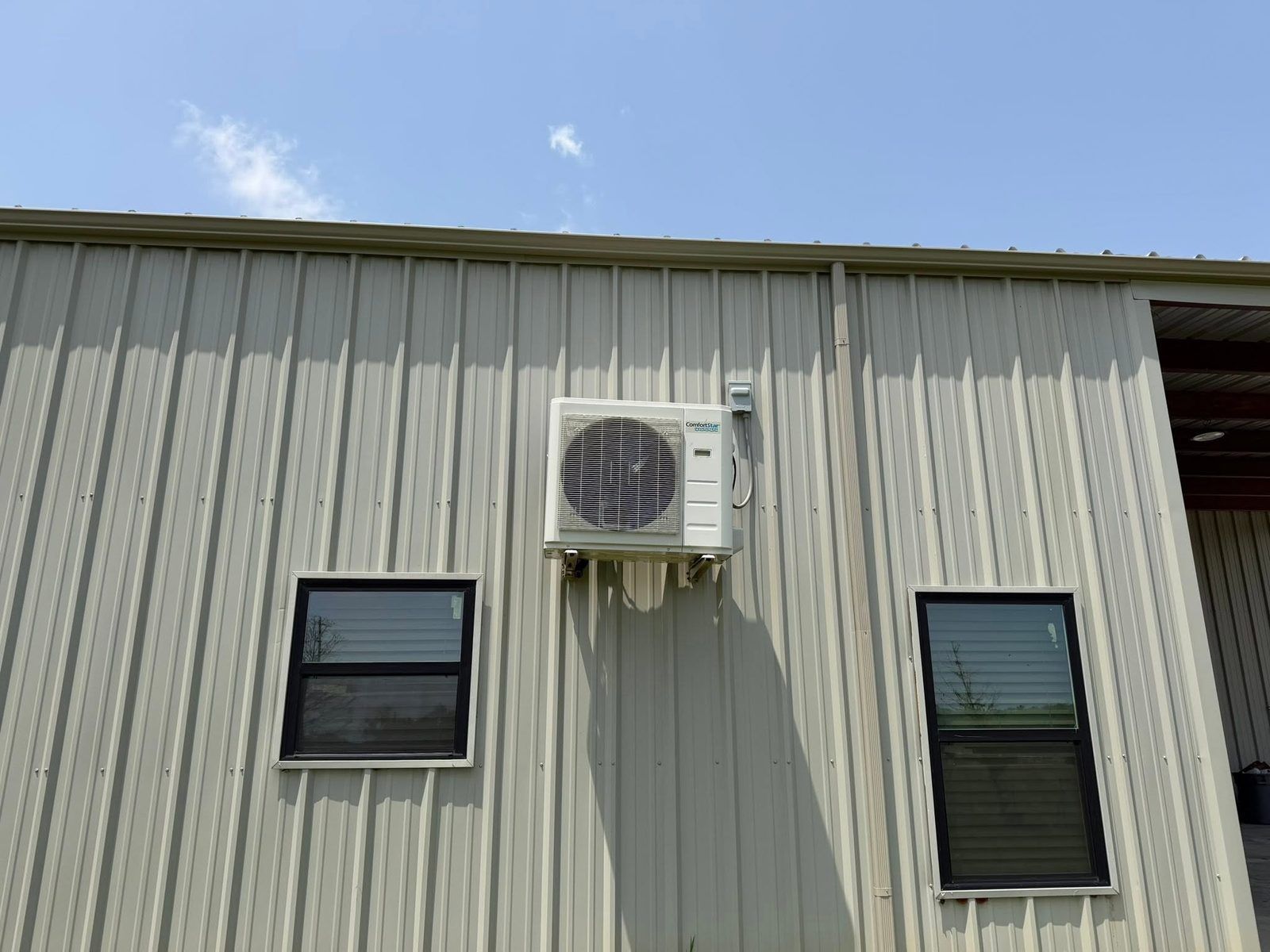Exterior of a beige metal building with three windows and a wall-mounted air conditioner under blue sky