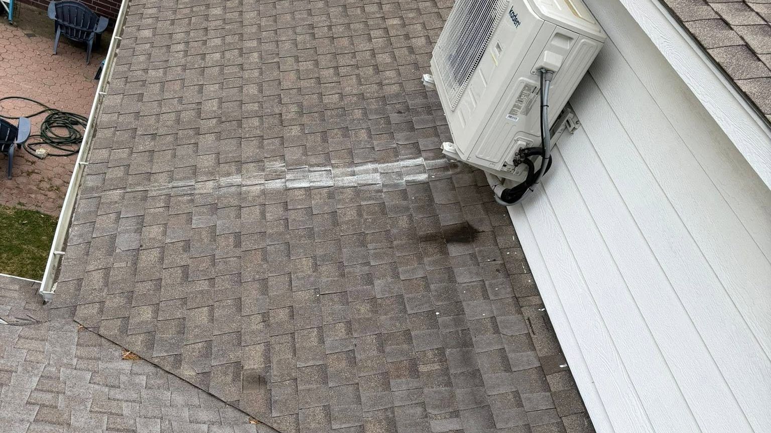 Roof shingles beside a house wall with an outdoor air conditioner unit and a dark stain on the roof
