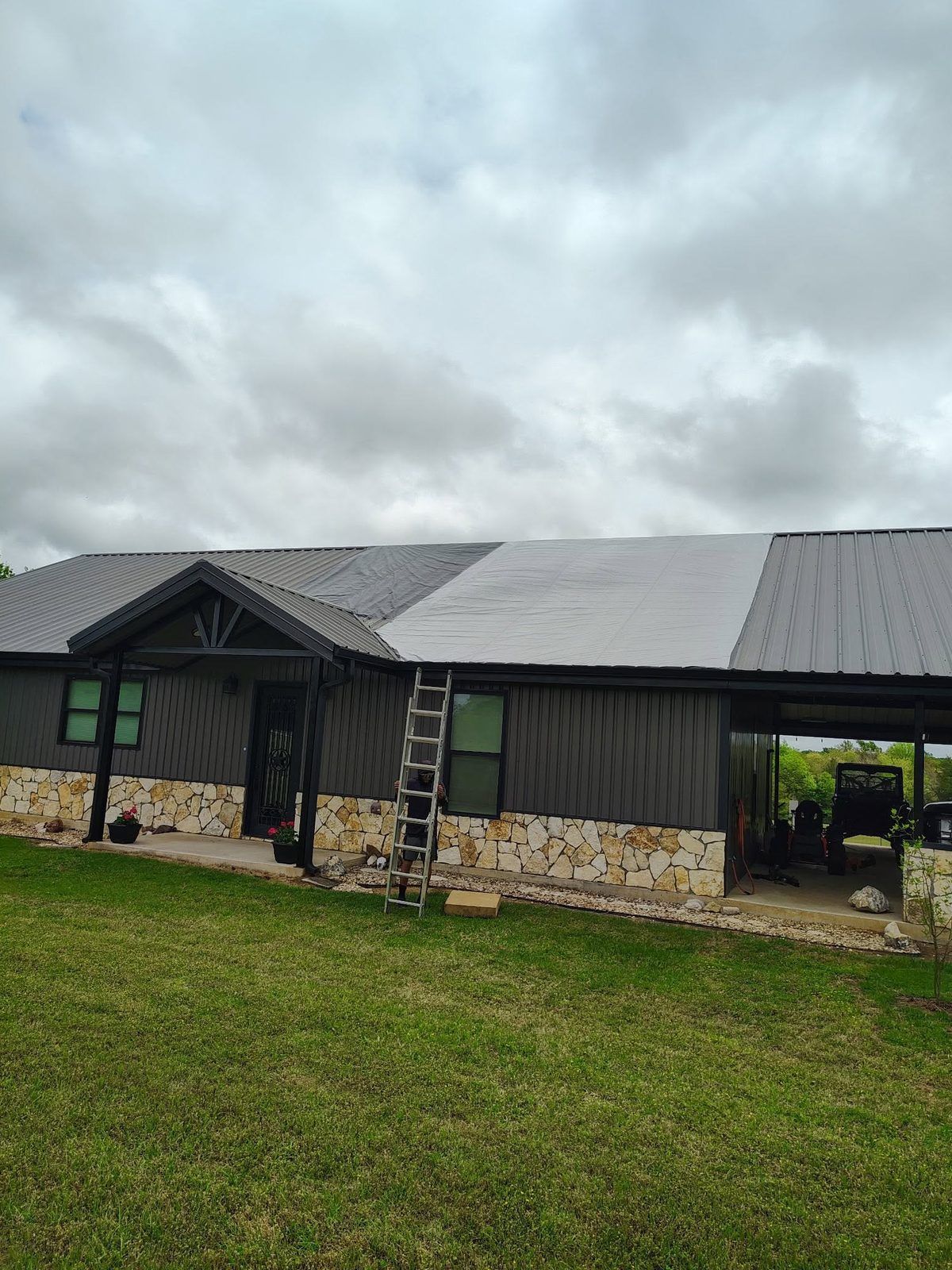 Long black house with stone base and ladder under cloudy sky, beside a green lawn