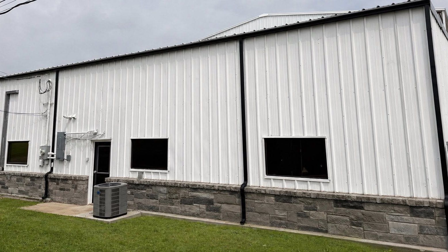 White metal industrial building with black window openings and stone foundation beside a grassy lot