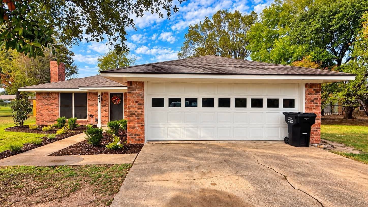 Single-story brick house with a white garage door and driveway, surrounded by trees and shrubs.