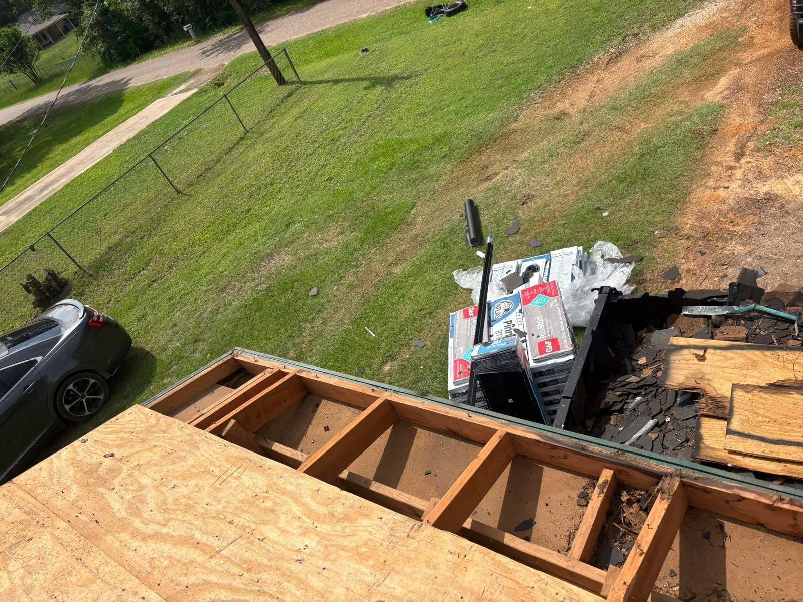 Roofing materials and exposed wooden framing on a sloped roof, with tools and debris below in a grassy yard.