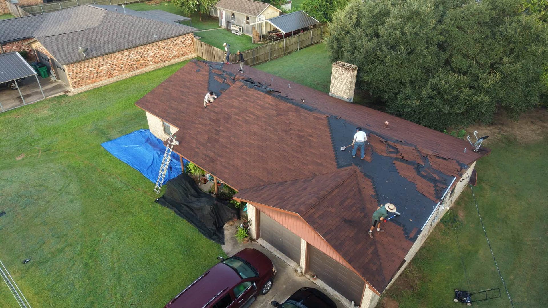 Aerial view of a house with a brown roof, blue tarp on one side, surrounded by a green lawn and parked car.