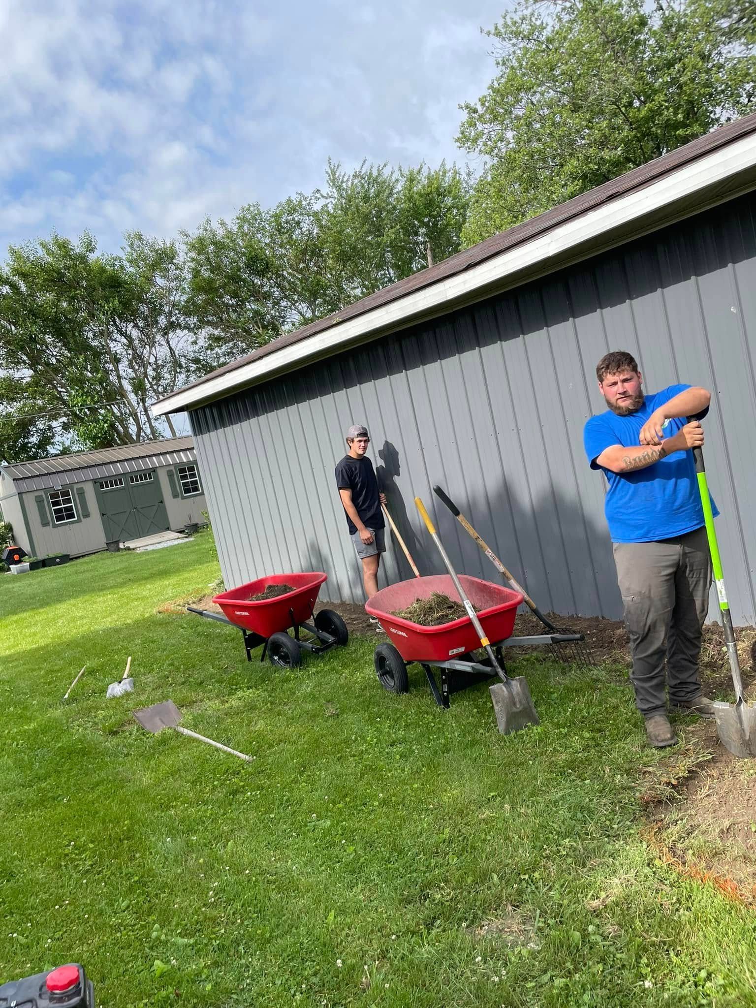Two men are working in a yard with wheelbarrows and shovels.