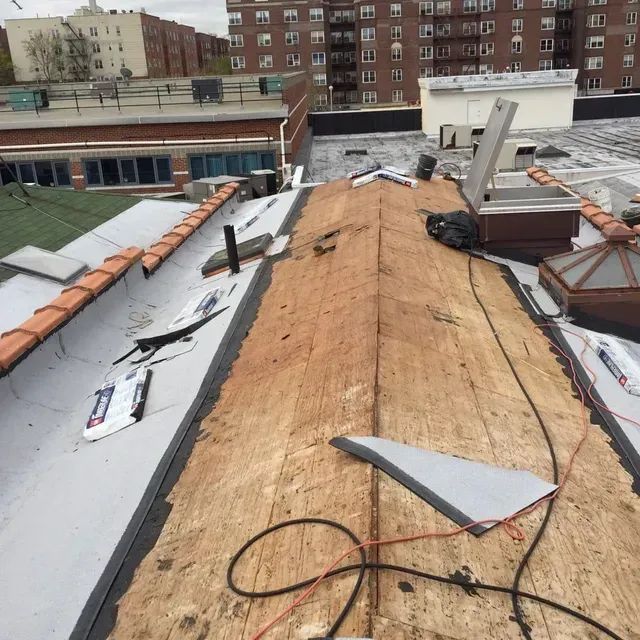 Rooftop with exposed wooden deck, partially covered with gray membrane. Skylight and buildings in background.