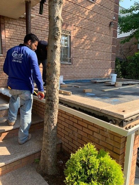 A man in a blue shirt is standing next to a tree in front of a brick building.