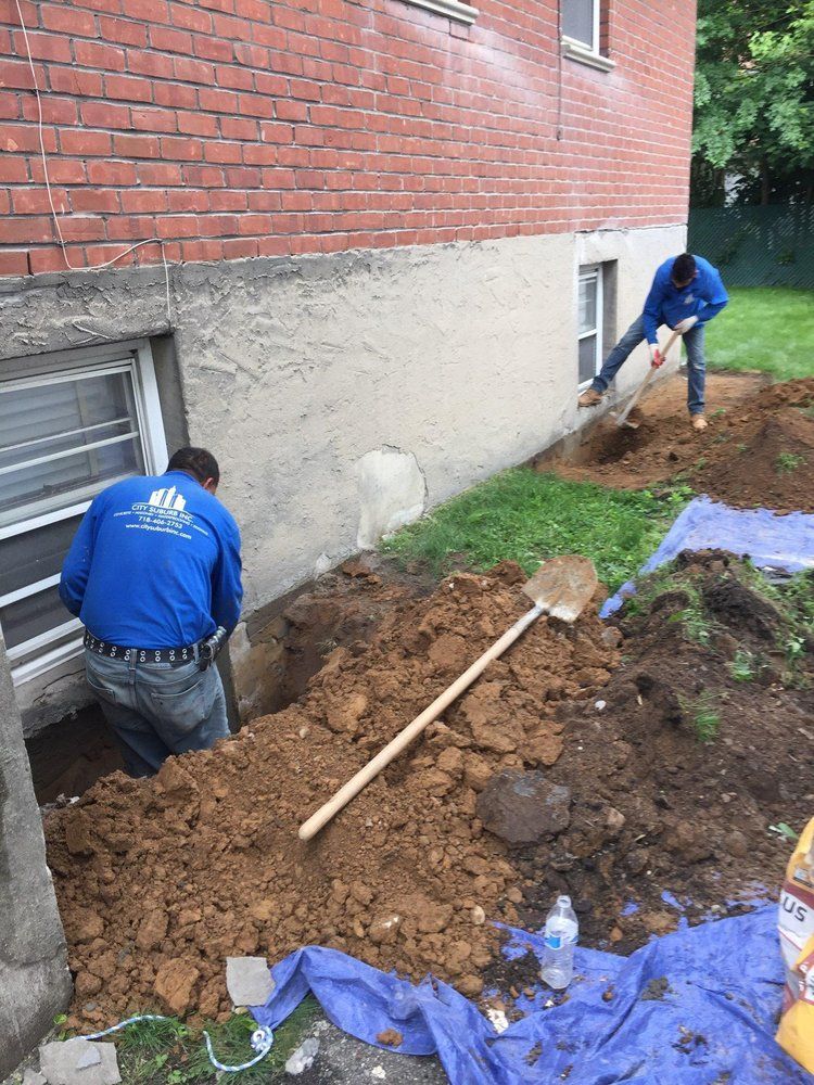 Two men are digging a hole in the ground in front of a brick building.