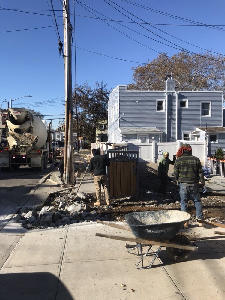 A man pushing a wheelbarrow on a sidewalk in front of a cement truck.