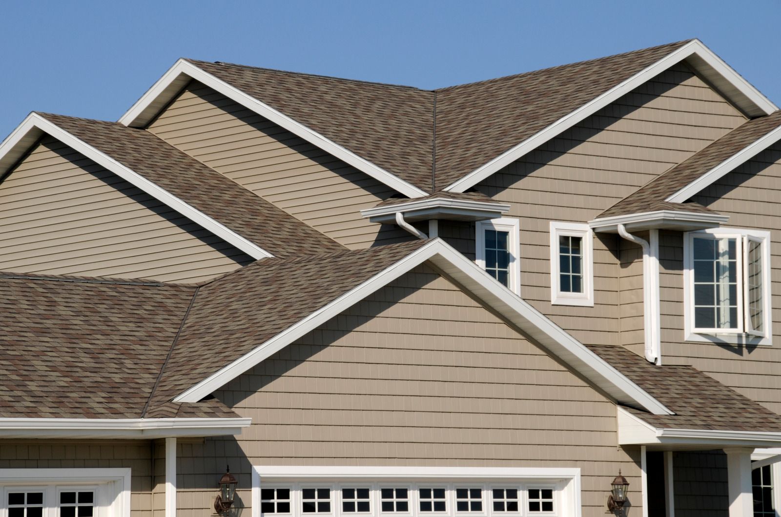 Tan house with brown shingled roof, white trim, and a garage door under a clear blue sky.