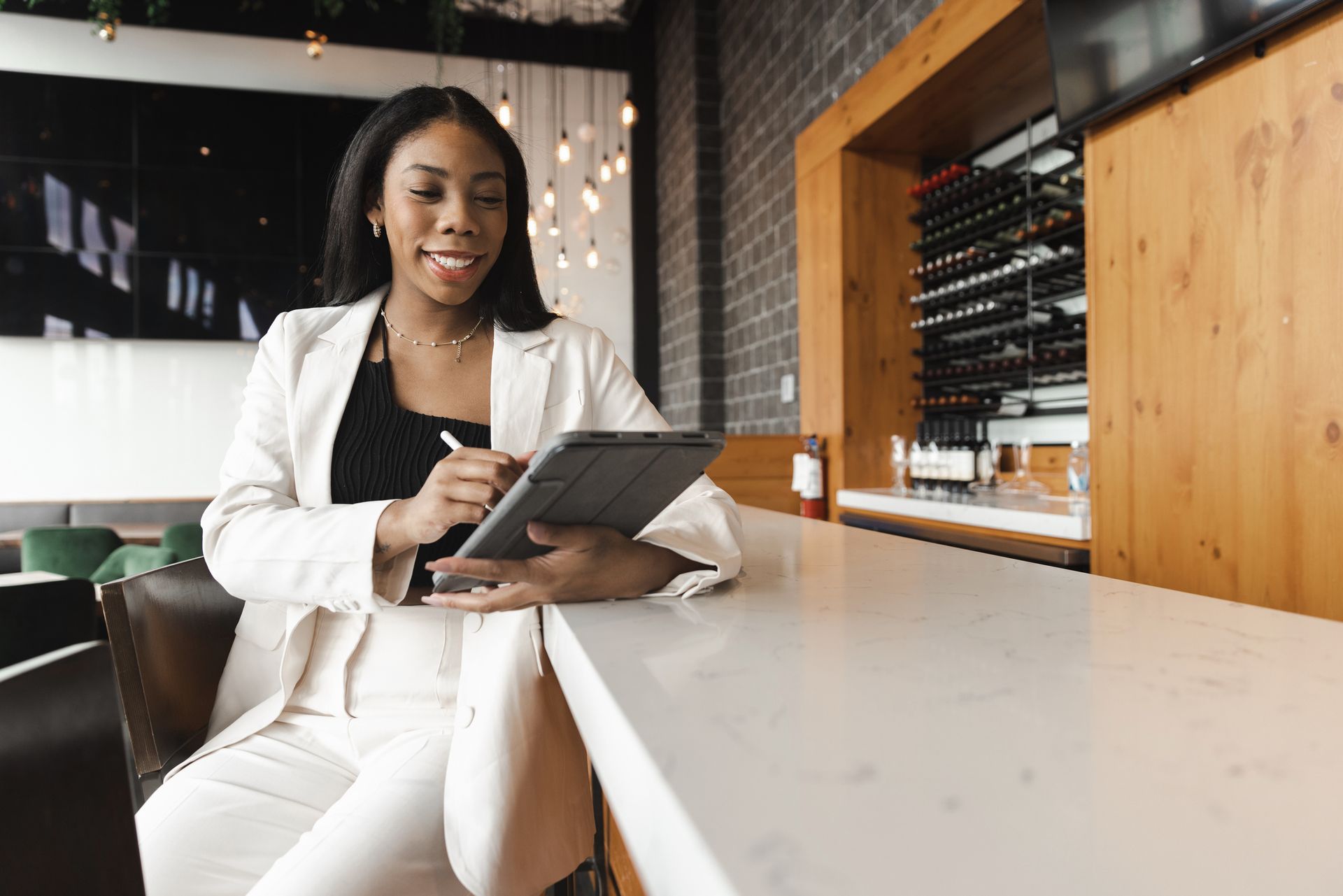 A Black woman in a cream suit, smiling, using a tablet with a stylus at a bar.
