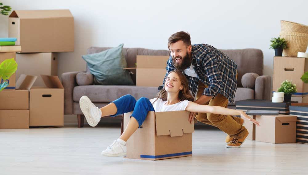 A man is holding a woman in a cardboard box in a living room.