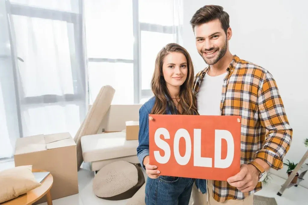 A man and a woman are holding a sold sign in their new home.