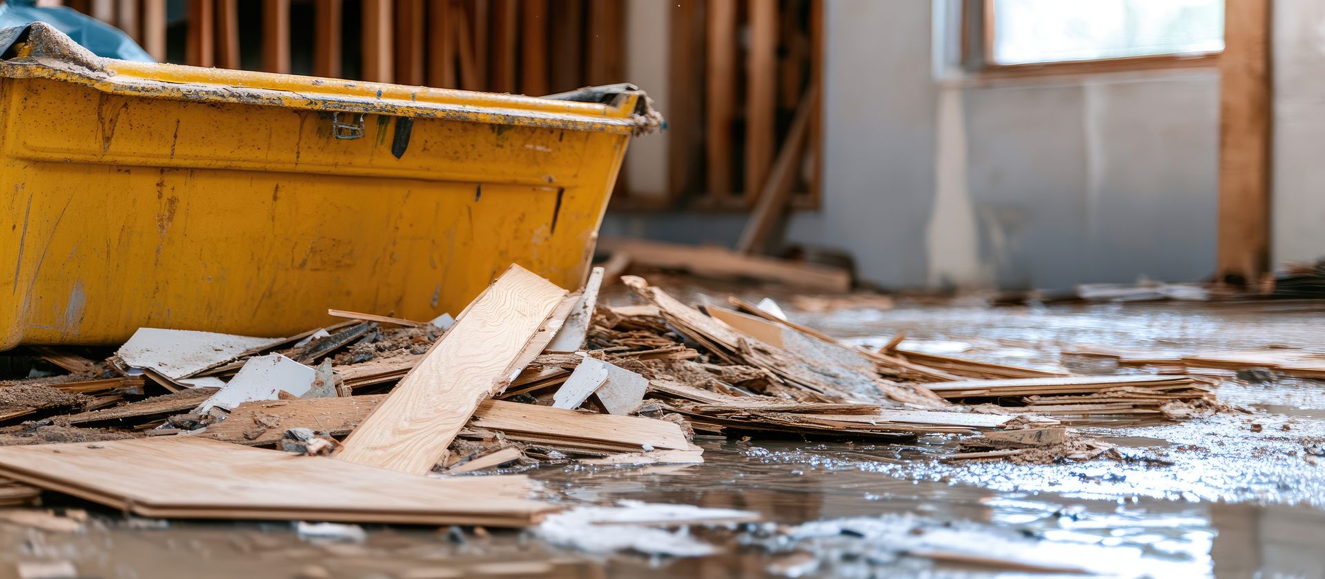A yellow bin sits amid scattered debris and standing water inside a room with exposed framing.