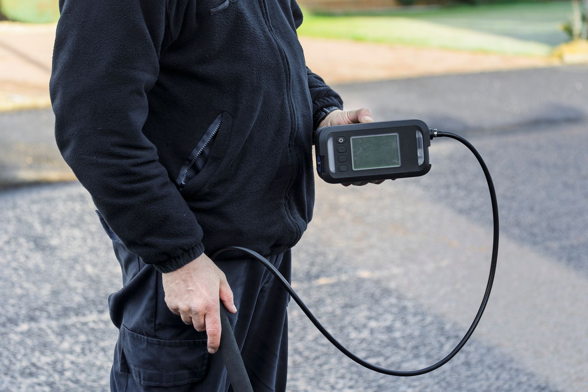 Person in black clothing holding a device with a screen, connected to a cable, possibly inspecting something outdoors.