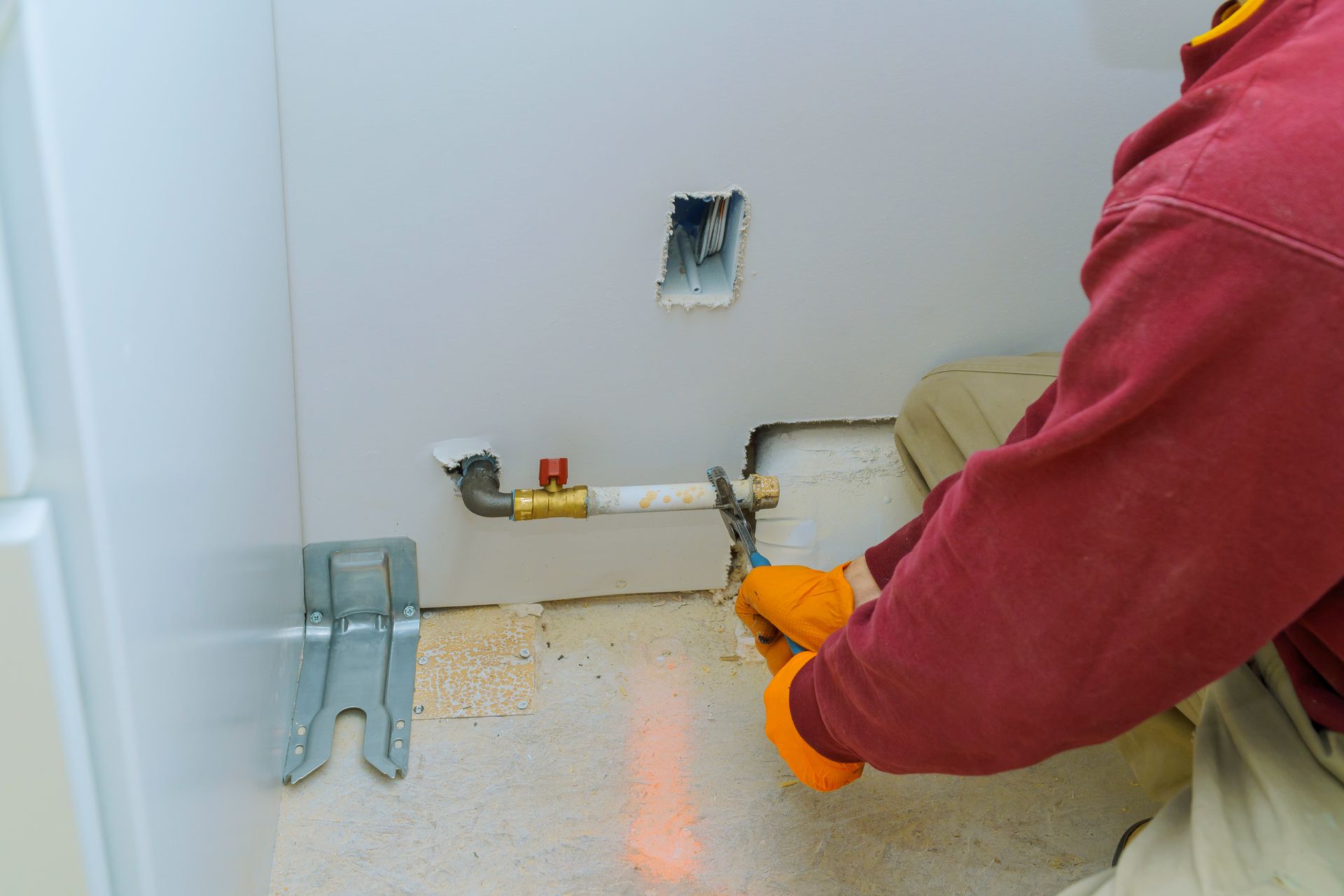 A person in orange gloves and a red shirt tightens a pipe with a wrench near a wall; a plumbing task.