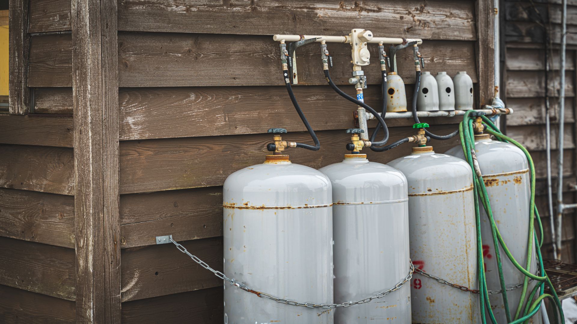 Gas cylinders secured with chains against a weathered wooden building. Hoses connect to a manifold.