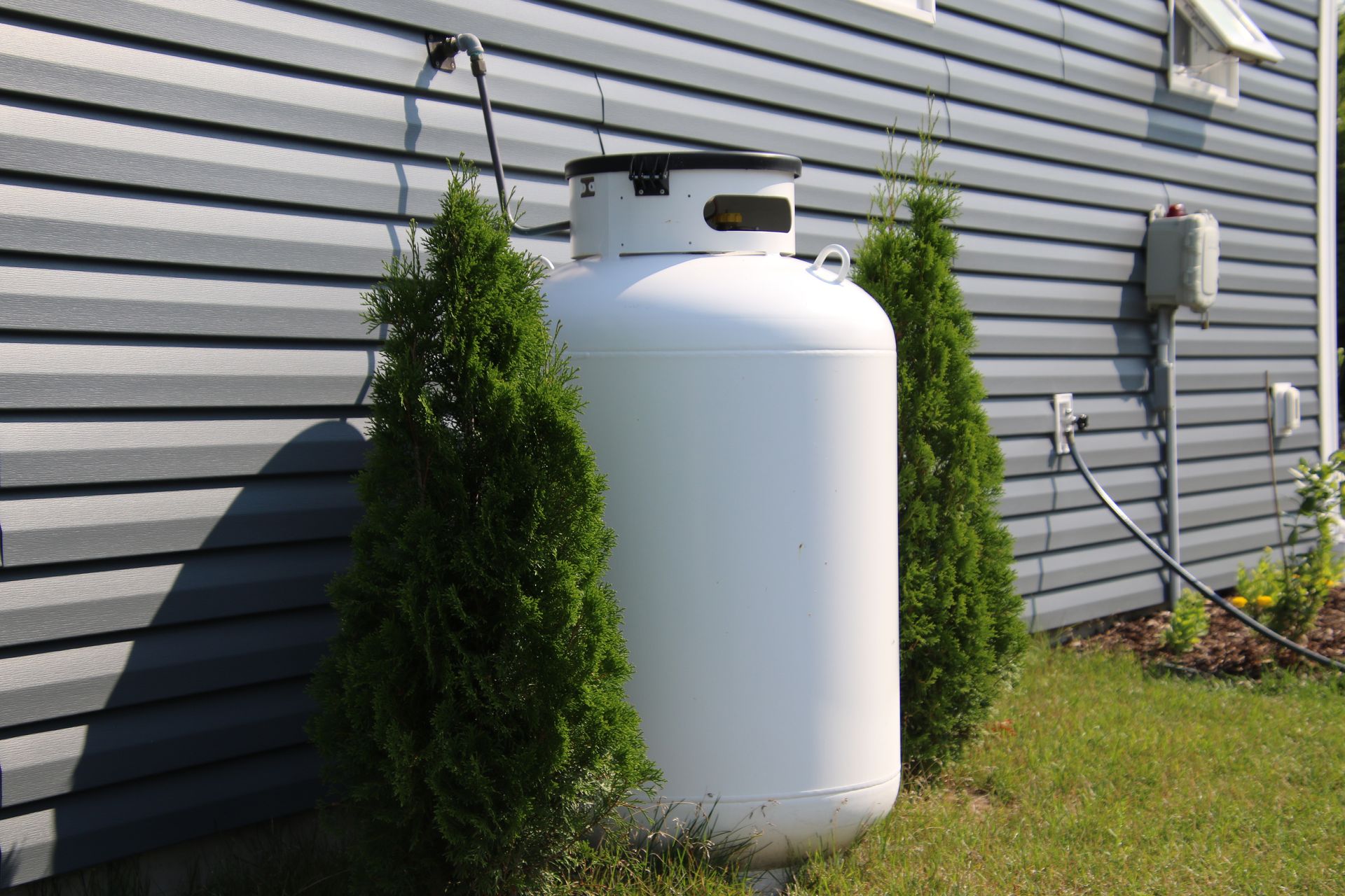 A large, white propane tank stands outside a building, flanked by two small evergreen trees.