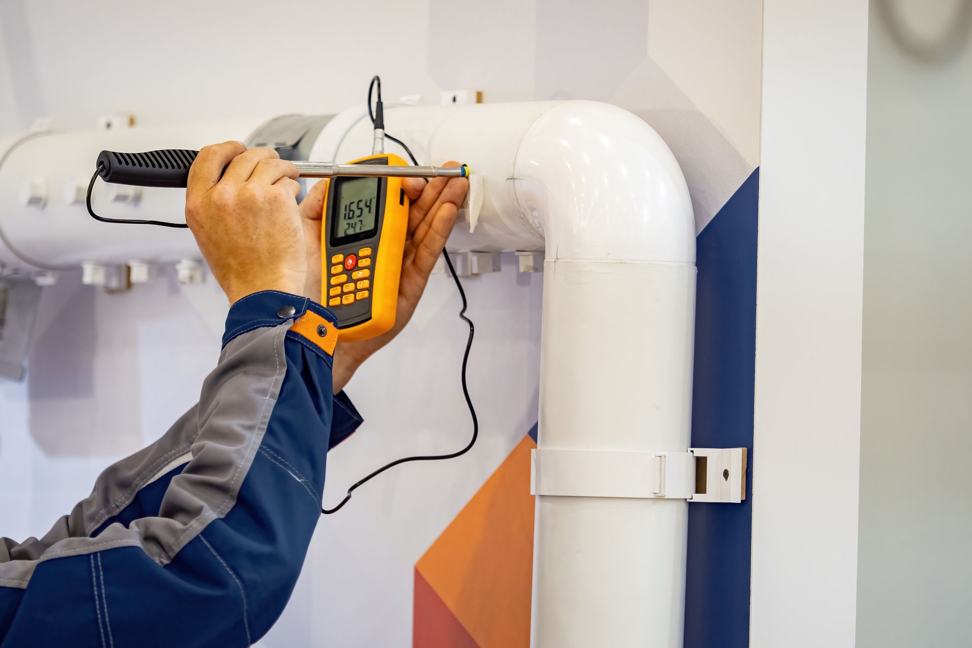 Person in work attire using a yellow meter to measure a white pipe in a facility setting.