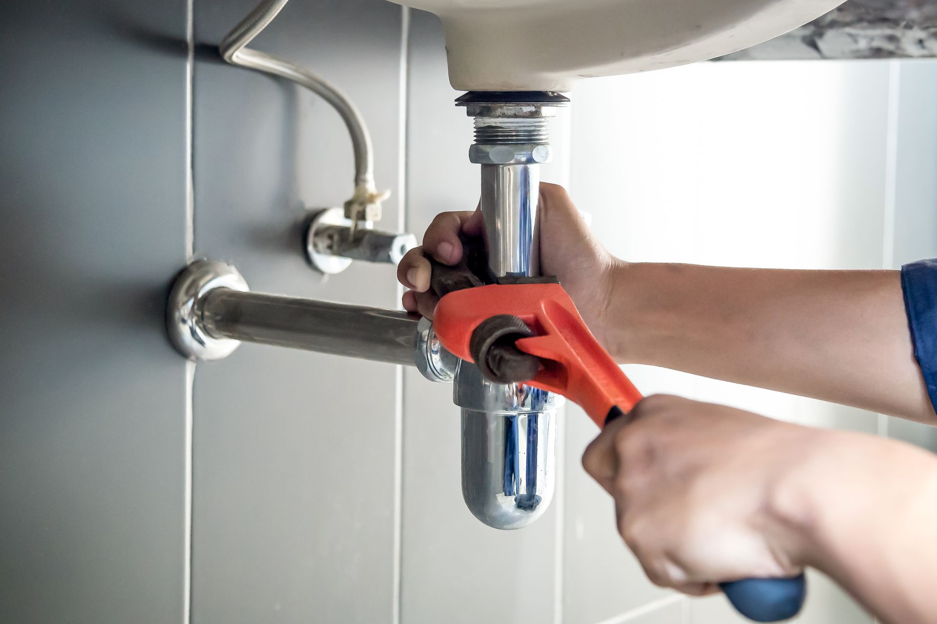 Person using a wrench to work on plumbing under a white sink. The pipes are chrome, and the wall is tiled.
