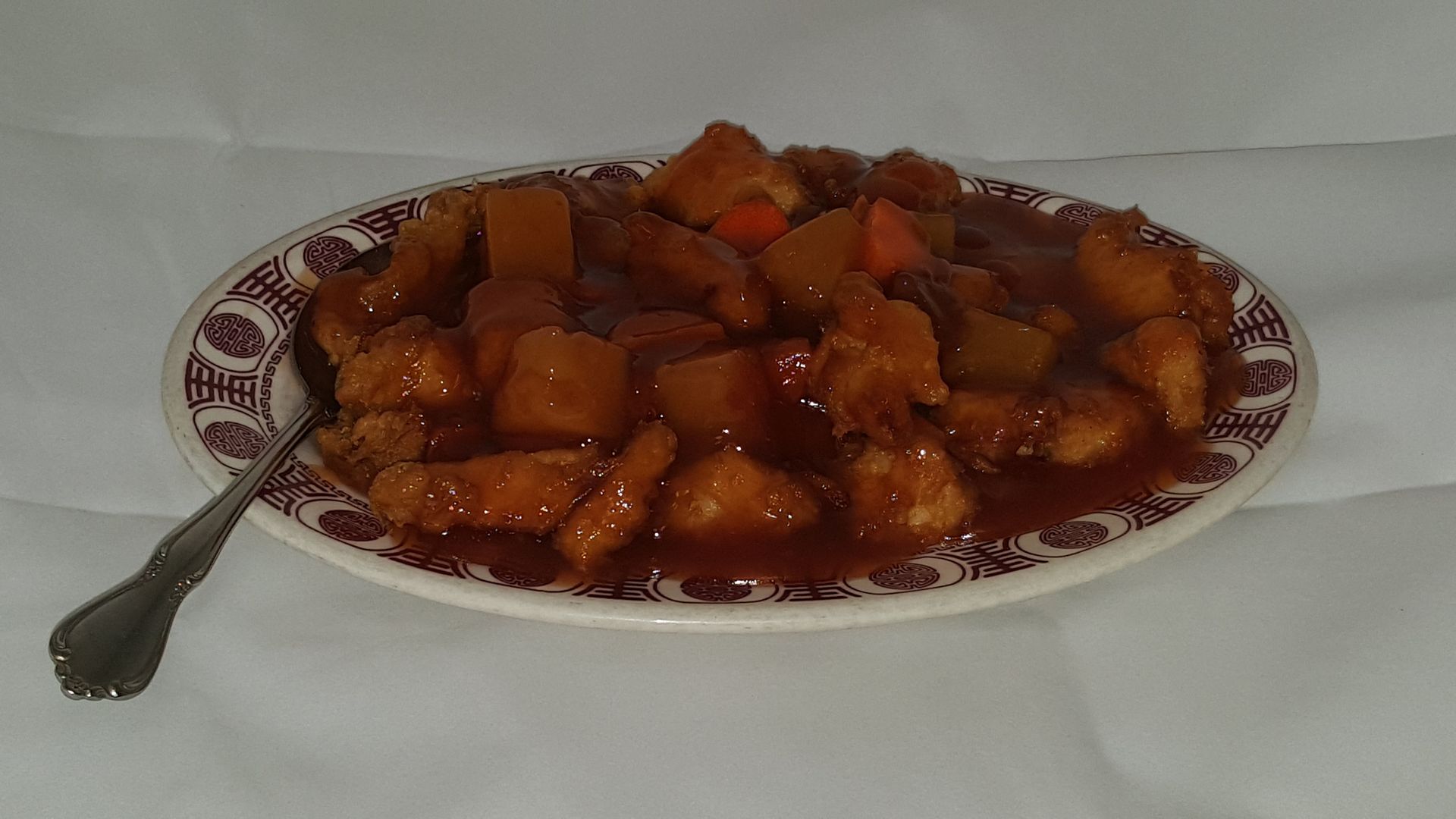 Sweet and sour chicken with pineapple and carrots in a red sauce, on a patterned plate with a spoon.