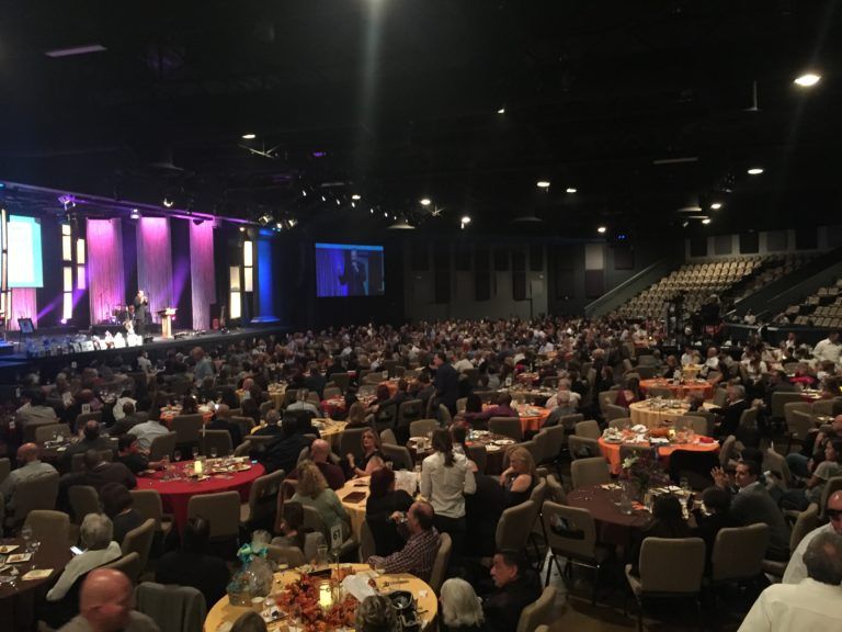 Large event in hall with audience at tables; stage at front with screens and presenters.