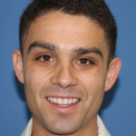 Man with a short haircut smiles at the camera, wearing a light-colored shirt, with a blue background.
