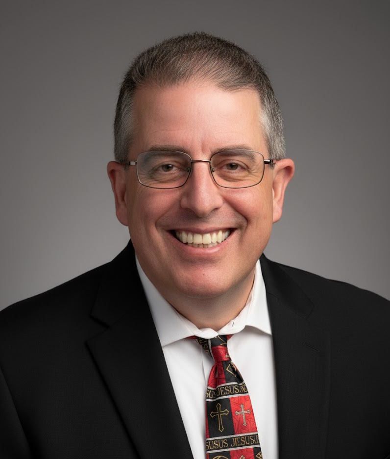 Man wearing glasses, smiling, in a suit and tie against a mottled gray background.