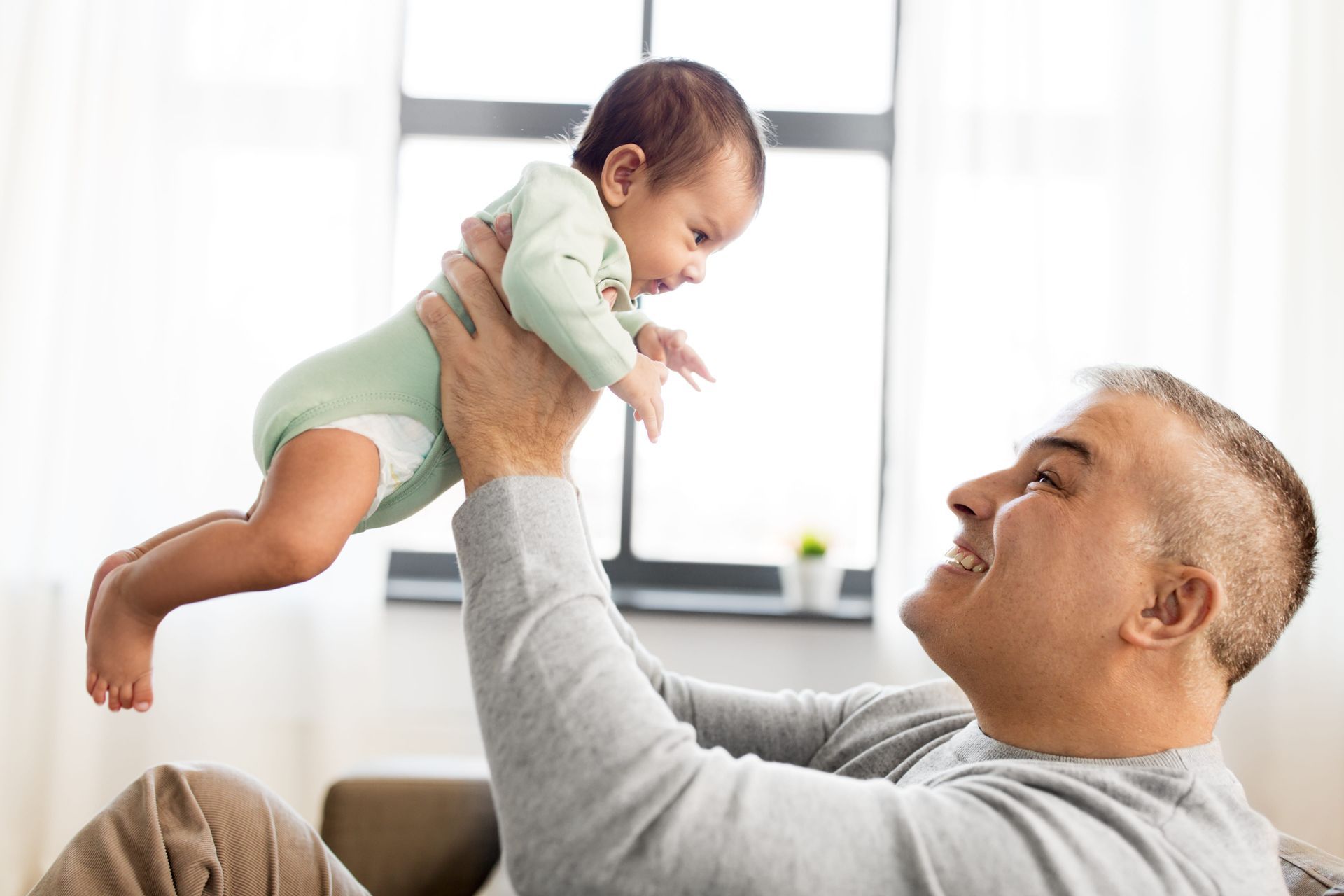 Adult holding smiling baby up indoors near a bright window