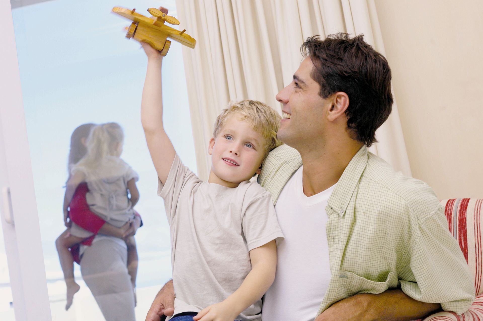 Boy holds toy airplane, looking at it with father. Family outside the window showing joint custody