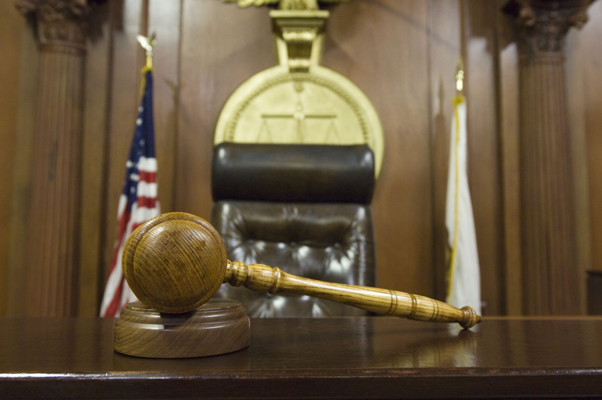 Gavel on judge's bench in courtroom with American and other flag; Justice scales emblem.