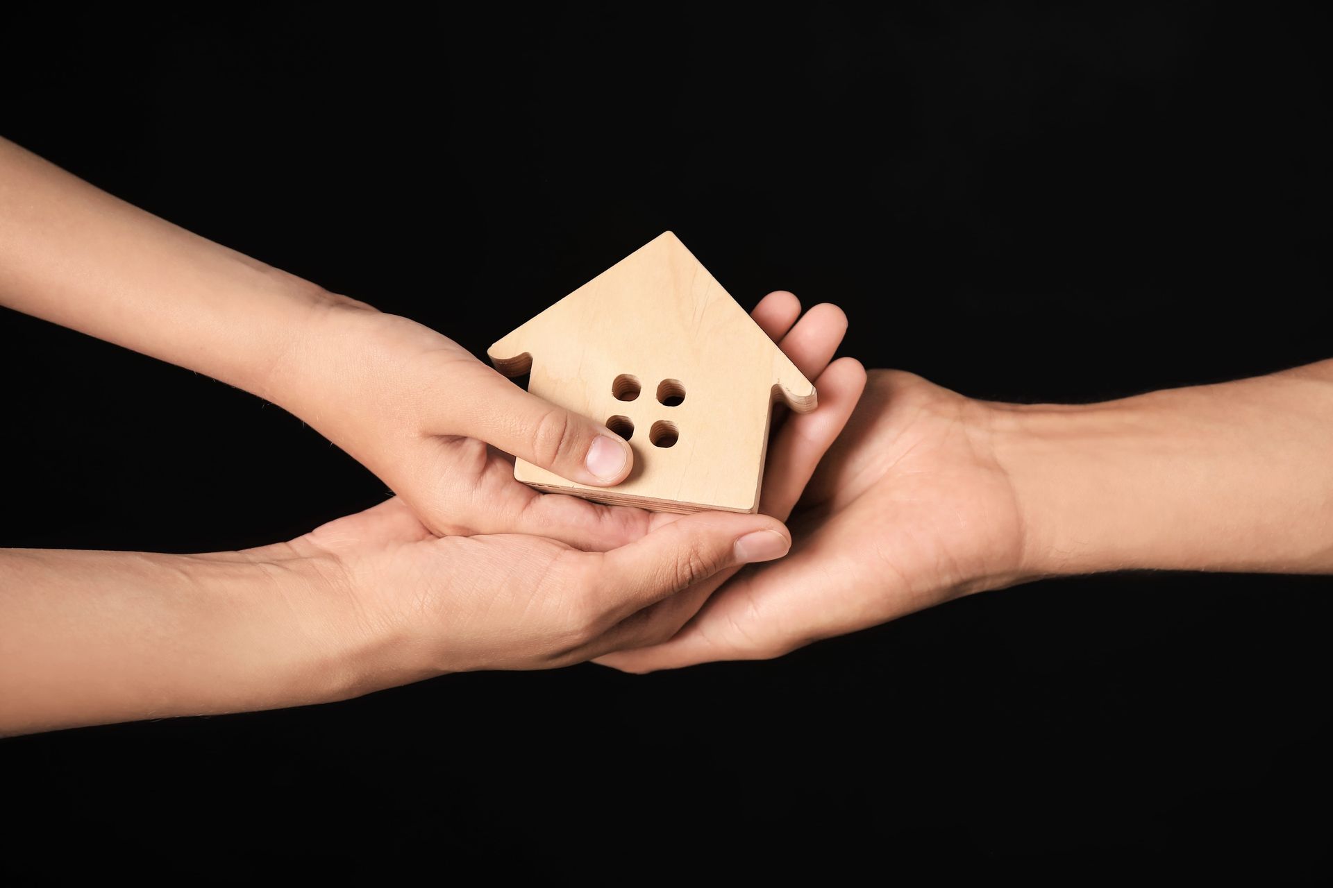 Hands passing a small wooden house over a black background, symbolizing support or home transfer
