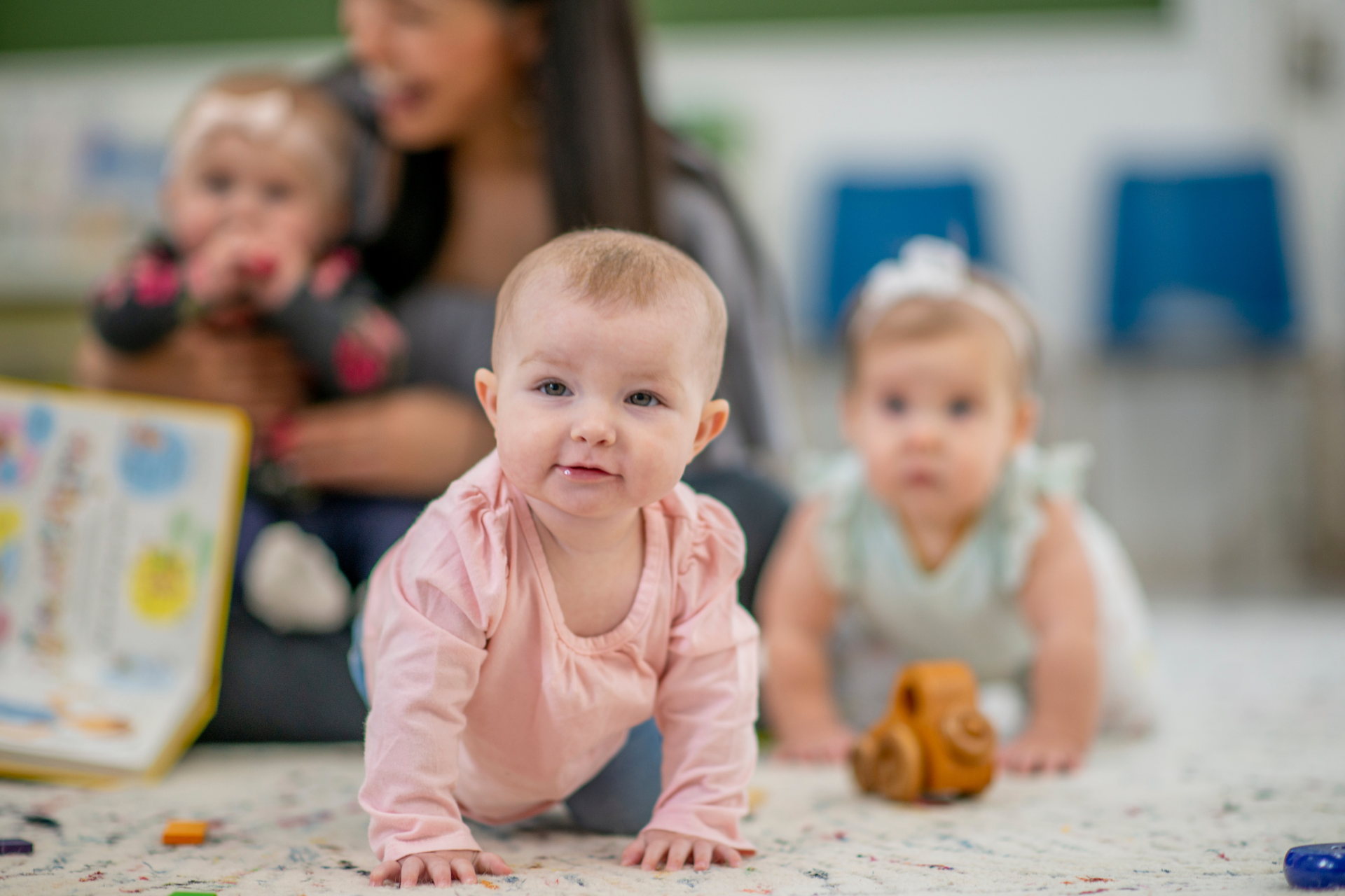 Babies crawling on a play mat in a daycare, one in pink in the foreground and another with a toy behind.