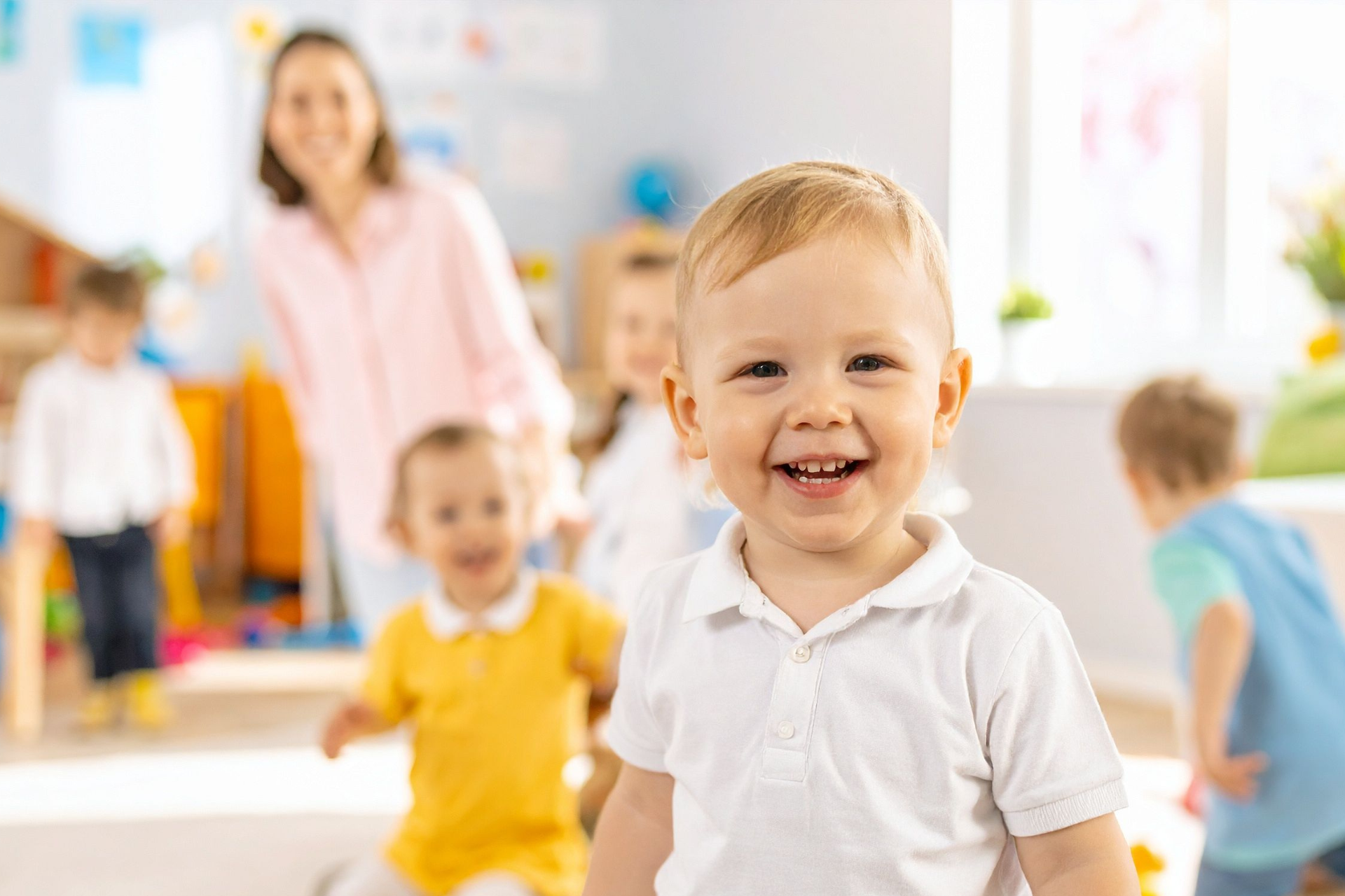Smiling toddler in white shirt at a colorful daycare with other children and a caregiver in the background