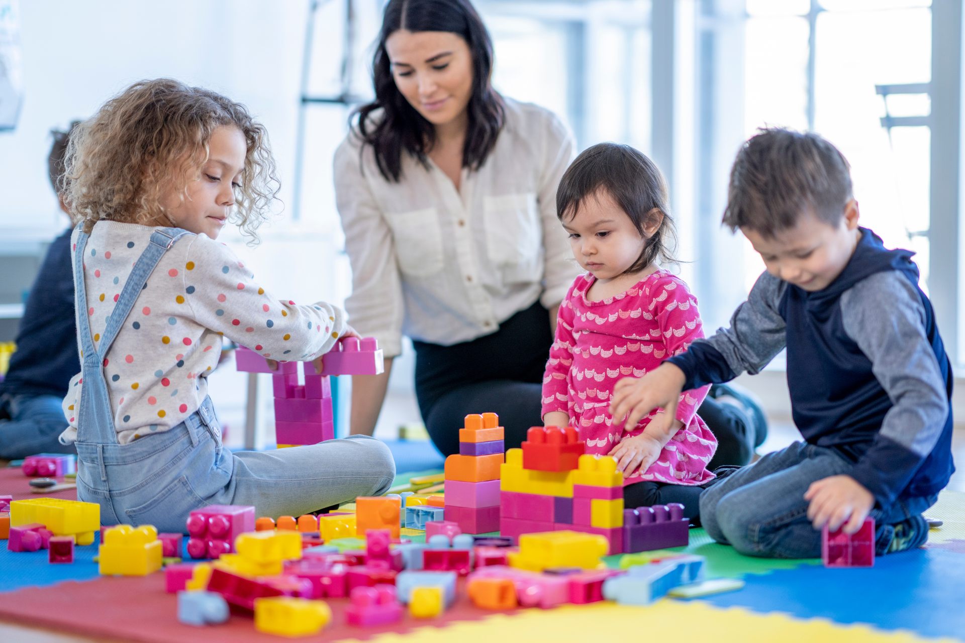 Children and a teacher playing with colorful building blocks on the floor in a bright classroom