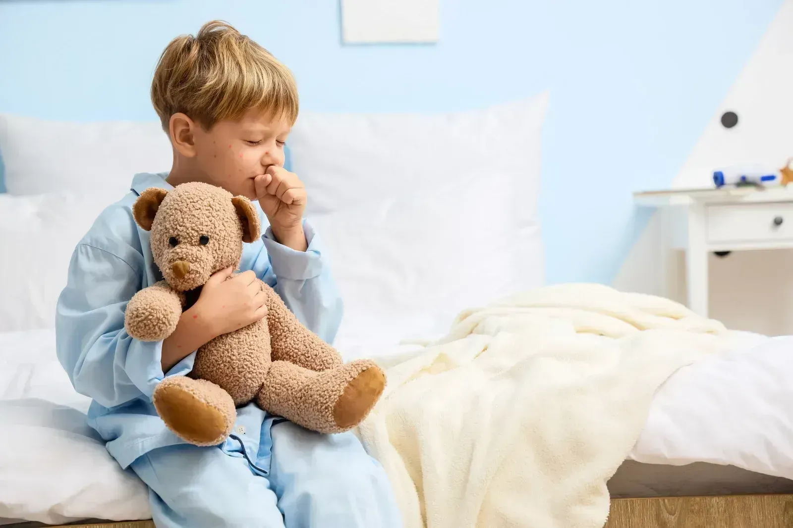 Young child sitting on a bed, hugging a teddy bear and looking thoughtful in a bright bedroom